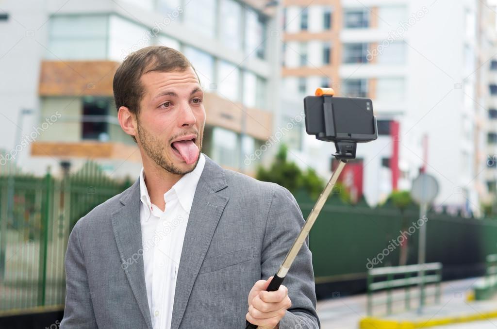 Man formal clothing posing with selfie stick in urban environment showing tongue