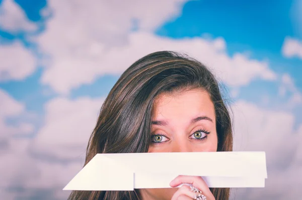 Beautiful brunette holding a paperplane covering her nose in front, cloud themed background ready to throw it — Stok fotoğraf