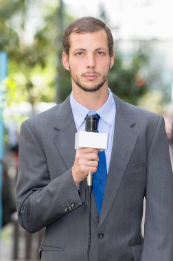 Attractive professional male news reporter wearing grey suit holding microphone, talking to camera from urban setting
