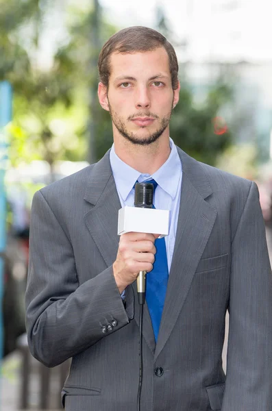 Attractive professional male news reporter wearing grey suit holding microphone, talking to camera from urban setting — Stockfoto