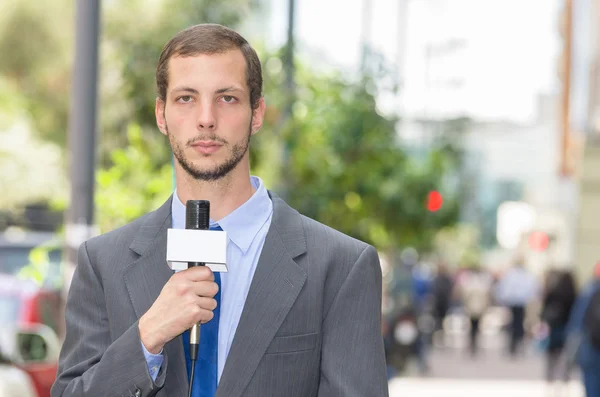 Attractive professional male news reporter wearing grey suit holding microphone, talking to camera from urban setting — Zdjęcie stockowe