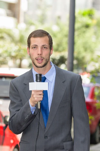 Attractive professional male news reporter wearing grey suit holding microphone, talking to camera from urban setting — Stock Photo, Image