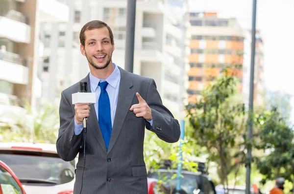 Attractive professional male news reporter wearing grey suit holding microphone, talking to camera from urban setting — 스톡 사진