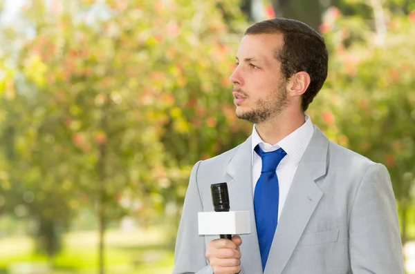 Successful handsome male news reporter wearing light grey suit working outdoors park environment holding microphone in live broadcasting — Stock Photo, Image