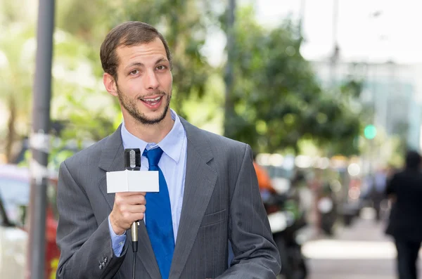 Attractive professional male news reporter wearing grey suit holding microphone, talking to camera from urban setting — Stock fotografie