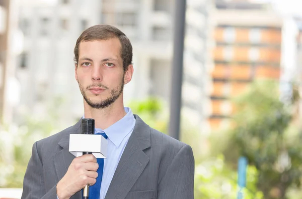 Atraente repórter profissional de notícias do sexo masculino vestindo terno cinza segurando microfone, falando com a câmera de ambiente urbano — Fotografia de Stock