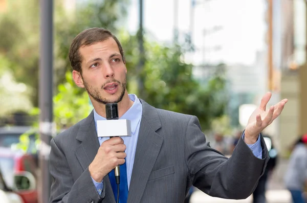 Atraente repórter profissional de notícias do sexo masculino vestindo terno cinza segurando microfone, falando com a câmera de ambiente urbano — Fotografia de Stock