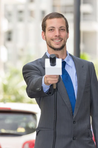 Attractive professional male news reporter wearing grey suit holding microphone, talking to camera from urban setting — 스톡 사진