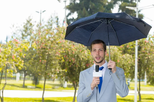 Successo bel giornalista maschio indossa tuta grigio chiaro che lavora in tempo piovoso all'aperto parco ambiente con microfono e ombrello, trasmissione in diretta — Foto Stock