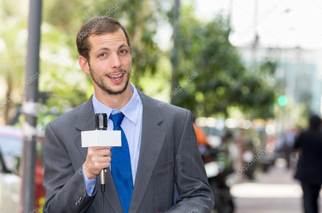 Attractive professional male news reporter wearing grey suit holding microphone, talking to camera from urban setting
