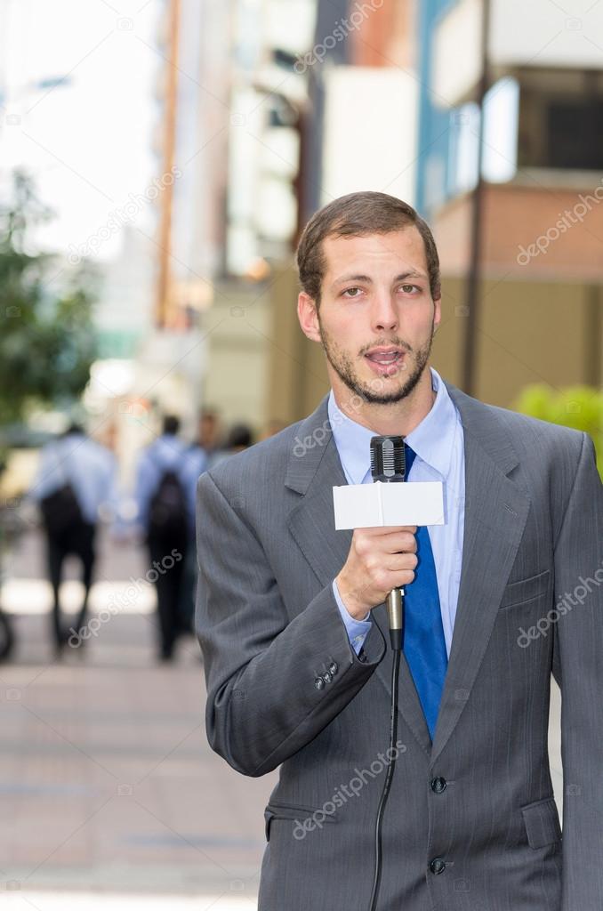 Attractive professional male news reporter wearing grey suit holding microphone, talking to camera from urban setting