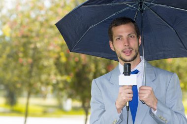 Successful handsome male journalist wearing light grey suit working in rainy weather outdoors park environment holding microphone and umbrella, live broadcasting clipart