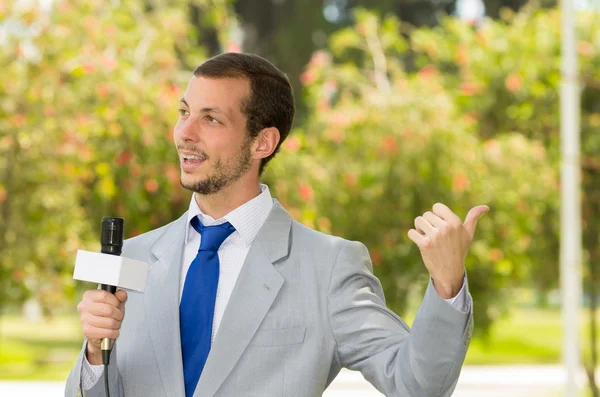 Successo bel reporter notizie maschile indossa tuta grigio chiaro che lavora all'aperto parco ambiente con microfono in diretta radiodiffusione — Foto Stock