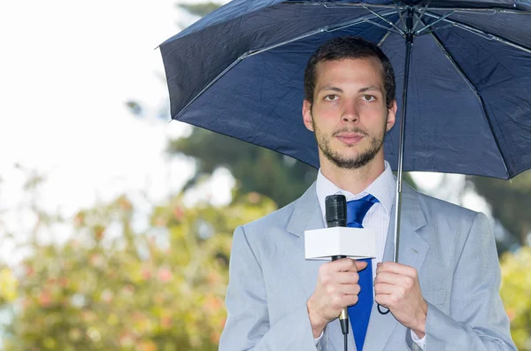 Jornalista bonito bem sucedido vestindo terno cinza claro trabalhando em clima chuvoso ao ar livre parque ambiente segurando microfone e guarda-chuva, transmissão ao vivo — Fotografia de Stock