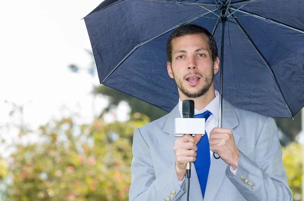 Un beau journaliste masculin portant un costume gris clair travaillant par temps pluvieux dans un environnement de parc avec microphone et parapluie, diffusion en direct — Photo