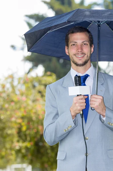 Jornalista bonito bem sucedido vestindo terno cinza claro trabalhando em clima chuvoso ao ar livre parque ambiente segurando microfone e guarda-chuva, transmissão ao vivo — Fotografia de Stock