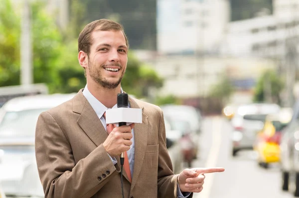 Successful attractive male journalist wearing brown suit working outdoors in traffic urban environment holding microphone, live broadcasting — Stock Photo, Image