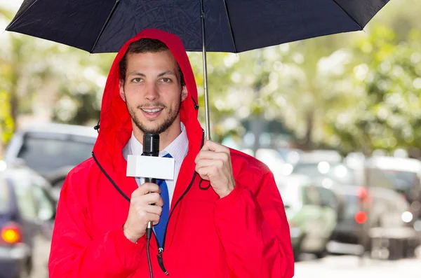 Periodista masculino guapo exitoso con chaqueta de lluvia roja que trabaja en clima lluvioso al aire libre entorno del parque sosteniendo micrófono y paraguas, transmisión en vivo — Foto de Stock
