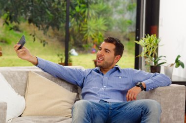 Hispanic man wearing jeans and blue shirt sitting in sofa posing for selfie smiling to cellphone camera