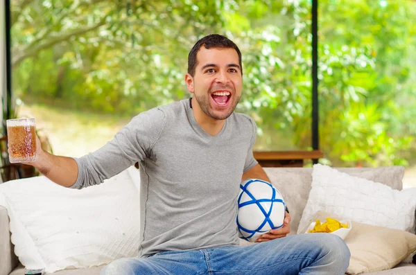 Латиноамериканец в джинсах в джинсах в сером свитере, сидящий на диване с энтузиазмом веселым выражением лица и смотрящий телевизор с пивом и футболом — стоковое фото