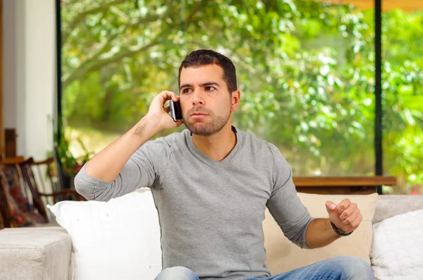 Hispanic man wearing denim jeans and grey sweater sitting in sofa leaning slightly forward while talking on phone seemingly with anticipation — Stock Photo, Image