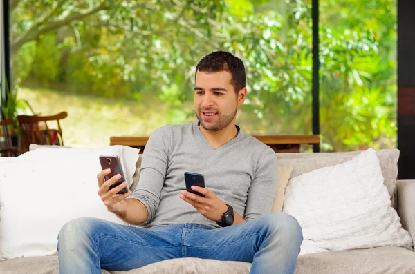 Hispanic man wearing denim jeans and grey sweater sitting on sofa comfortably with a two cellphones, one in each hand — Stock Photo, Image