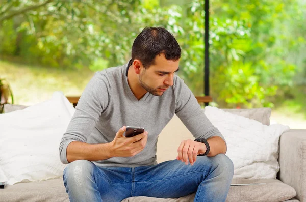 Hispanic man wearing denim jeans with grey sweater sitting in sofa holding cellphone and looking at the watch on his left arm — Stock Photo, Image