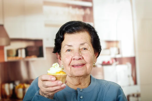 Older hispanic happy woman wearing blue sweater sitting in front of camera showing off cupcake with cream topping — Stok fotoğraf