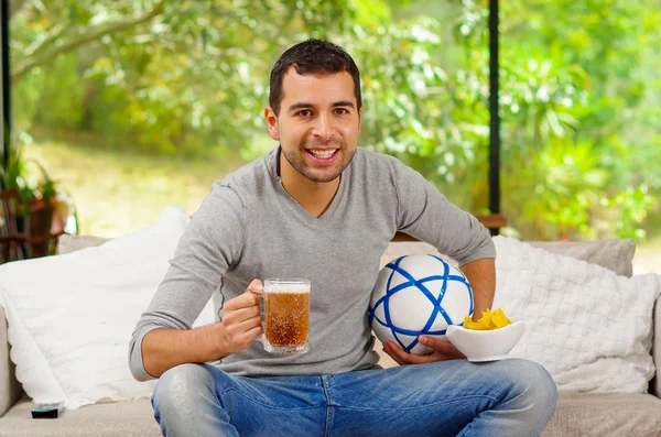 Hombre hispano vistiendo vaqueros con suéter gris sentado en un sofá viendo con entusiasmo la televisión sosteniendo cerveza y fútbol — Foto de Stock