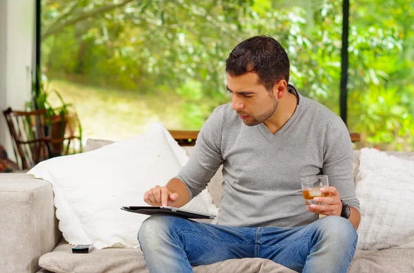 Hispanic man wearing denim jeans and grey sweater sitting in sofa with tablet resting on knee looking at screen holding glass of dark colored beverage — ストック写真