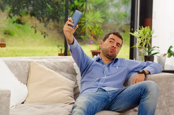 Hispanic man wearing jeans and blue shirt sitting in sofa posing for selfie making kiss lips leaning backwards — Stock Photo, Image