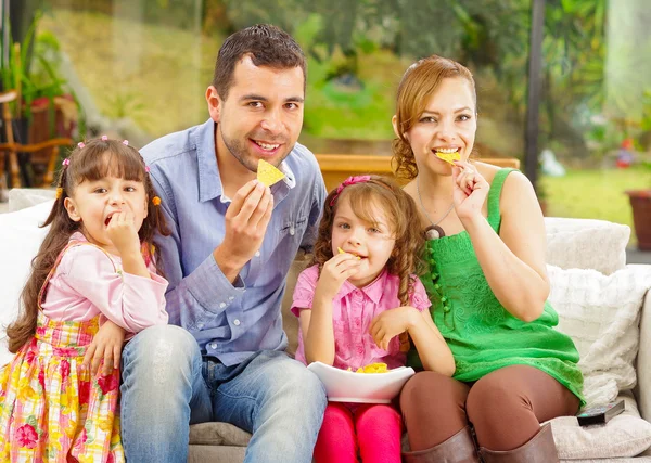 Retrato familiar de padre, madre y dos hijas sentadas juntas en un sofá disfrutando de unos nachos felizmente sonriendo a la cámara — Foto de Stock