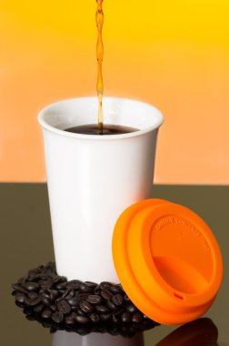 Great image showing stream of coffee pouring into white cup from above with coffe beans on surface sourrounding mug and orange lid sunset colored background