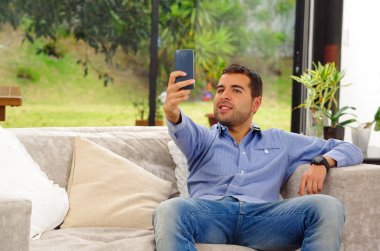Hispanic man wearing jeans and blue shirt sitting in sofa posing for selfie holding cellphone carefully smiling
