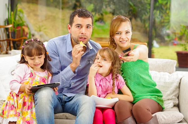 Family portrait of father, mother and two daughters sitting together in sofa playing with tablet while enjoying some nachos happily smiling to camera. — Stock fotografie