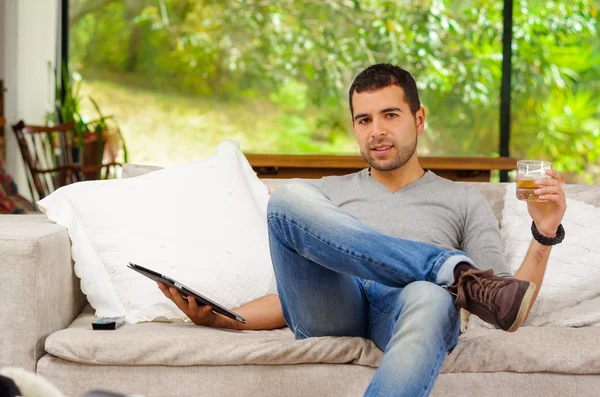 Hispanic man wearing denim jeans and grey sweater sitting on sofa laidback position holding a tablet with right hand, dark colored beverage glass in left looking at camera — Zdjęcie stockowe