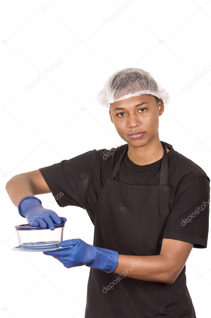 Hispanic young man wearing blue cleaning gloves and plastic shower cap securing lid on circular pyrex looking to camera