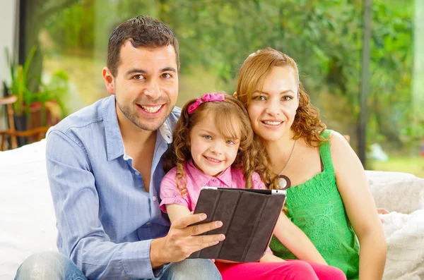 Family portrait of father, mother and daughter sitting together in sofa holding tablet looking towards camera — Stockfoto