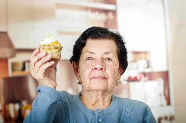 Older hispanic woman wearing blue sweater sitting in front of camera holding up a yellow cupcake with cream topping — Stock fotografie