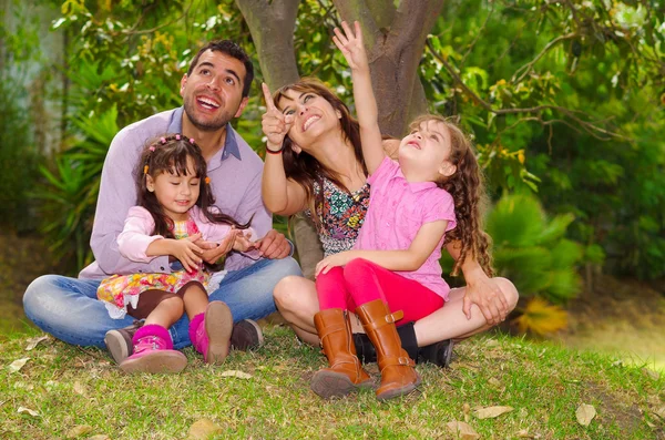 Family portrait of father, mother and two daughters sitting together in garden environment pointing looking upwards towards the sky — Stock Photo, Image