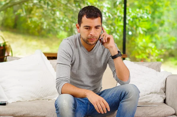 Hispanic man wearing denim jeans with grey sweater sitting in sofa leaning slightly forward while talking on phone and facial expression as paying attention — Zdjęcie stockowe