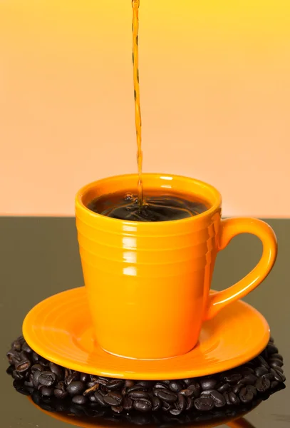 Great image showing stream of coffee pouring into orange cup from above with mug sitting on coffe beans and shiny surface, sunset colored background — Stock Photo, Image