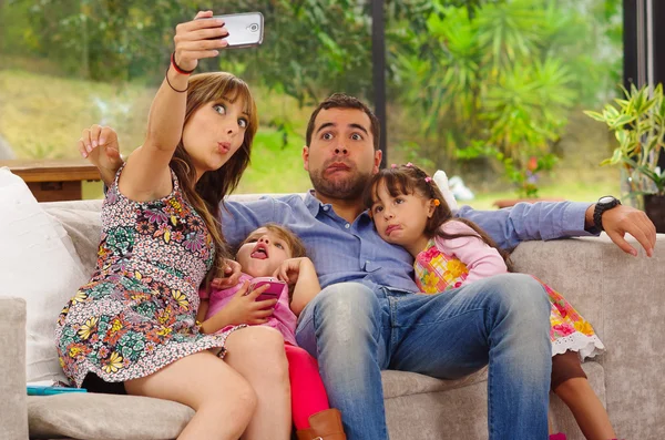 Retrato familiar de padre, madre y dos hijas sentadas juntas en sofá posando para selfie haciendo caras graciosas —  Fotos de Stock