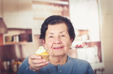 Older hispanic happy woman wearing blue sweater sitting in front of camera showing off cupcake with cream topping