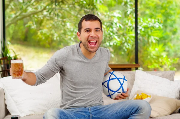 Hombre hispano vistiendo vaqueros con suéter gris sentado en el sofá con entusiasmo alegre expresión facial viendo la televisión sosteniendo cerveza y fútbol — Foto de Stock