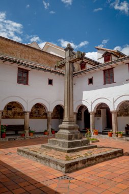 Large stone cross located inside courtyard of San Diego church, beautiful colonial white building stretching around the small plaza covered in red tiles