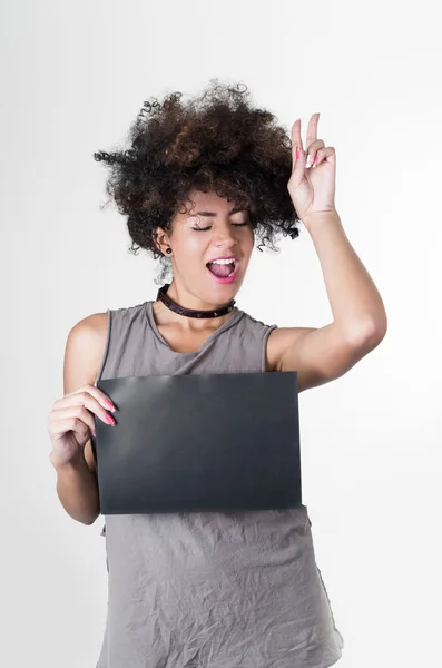 Hispanic brunette rebel model with afro like hair wearing grey sleeveless shirt holding blank board as posing for mugshot concept, making peace symbol using left hand up in air next to head — Stockfoto
