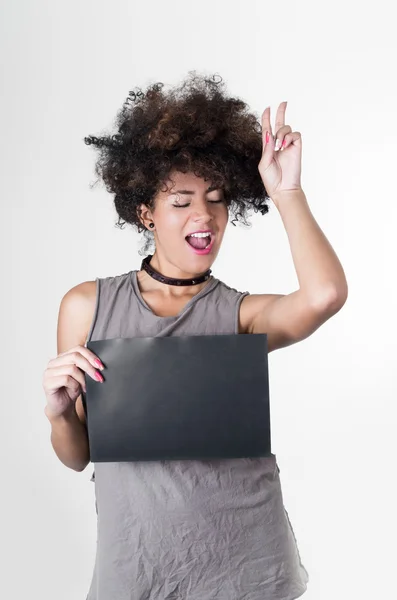 Modelo rebelde morena hispânica com afro como cabelo vestindo camisa cinza sem mangas segurando placa em branco como posando para conceito mugshot, fazendo símbolo de paz usando a mão esquerda para cima no ar ao lado da cabeça — Fotografia de Stock