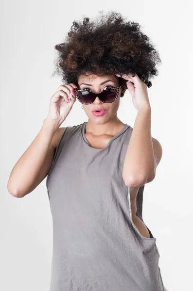 Modelo rebelde morena hispana con afro como el pelo usando camisa gris sin mangas poniéndose gafas de sol y mirando a la cámara —  Fotos de Stock