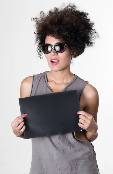 Hispanische brünette Rebellin mit afro-artigen Haaren, grauem ärmellosen Hemd und Sonnenbrille, die ein leeres Brett hochhält, um für das Mugshot-Konzept zu posieren — Stockfoto